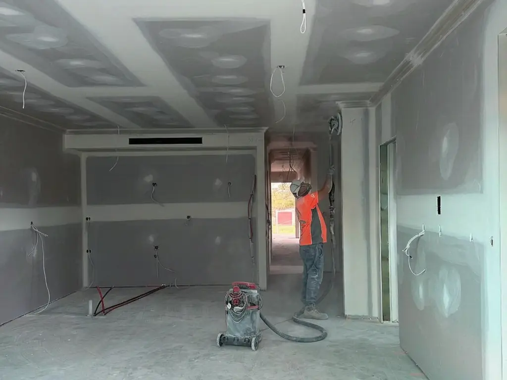 Sanding a set ceiling with a dust extracted sander in an open plan living space, the last pass before a Level 5 finish is signed off.