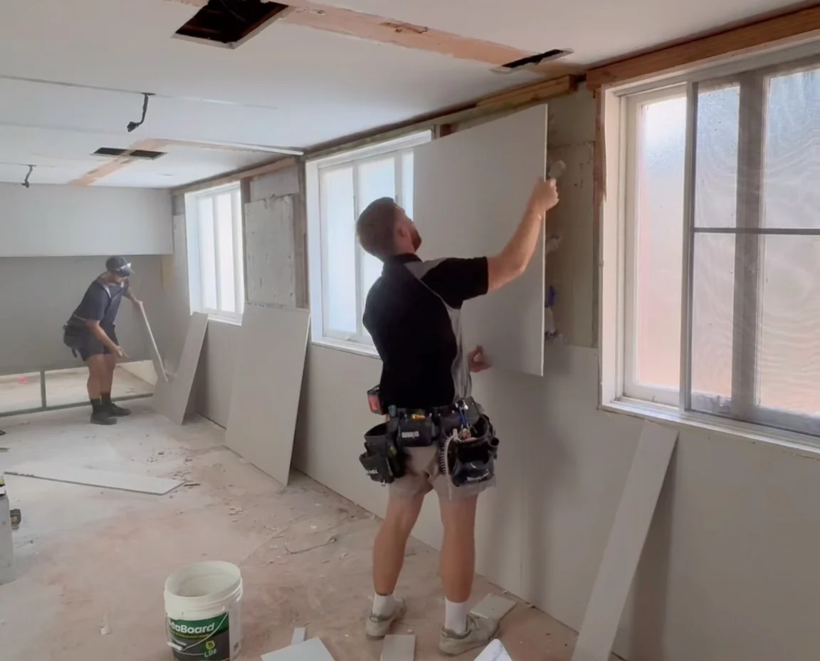 Two plasterers lining a room in a Sydney home being renovated, one holding a sheet of plasterboard against the wall frame under a window while the other cuts a sheet behind him, with the ceiling already sheeted and more sheets stacked against the wall.