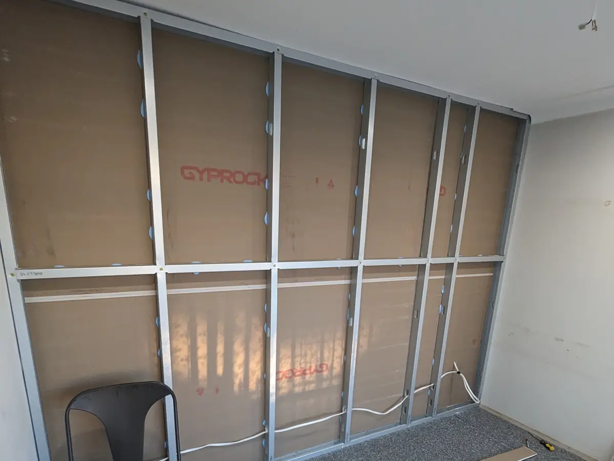 A new steel stud wall framed up inside an existing carpeted room, with Gyprock sheets stacked on the floor ready to hang.