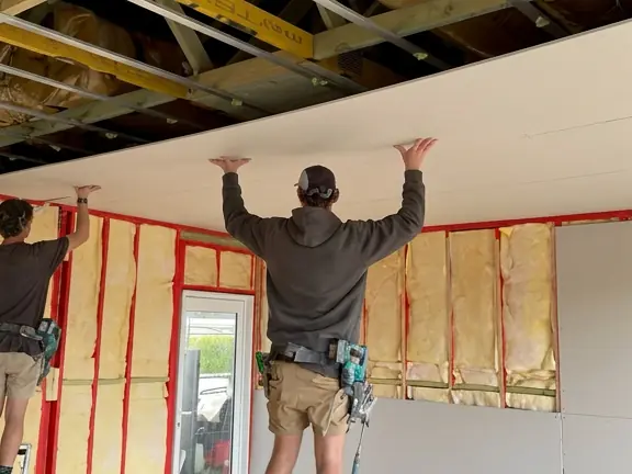 Two of us holding a sheet up to the ceiling while the walls stand packed with acoustic insulation batts between the studs, and more insulation sitting between the ceiling joists above.