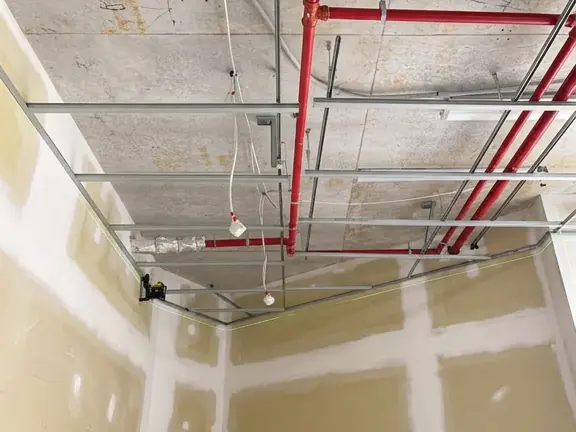 Looking up into a suspended ceiling frame before the sheets go on, with the sprinkler main and the services running above the grid.